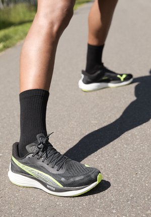 Leichte schwarze Laufschuhe mit strukturiertem Mesh-Obermaterial, grünen Akzenten und weißer Sohle. Getragen mit schwarzen Knöchelsocken auf einem gepflasterten Weg.