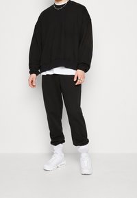 Černá oversized mikina s kleslými rameny, přes kterou je vrstvená bílá košile. V kombinaci s odpovídajícími černými tepláky a bílými teniskami.