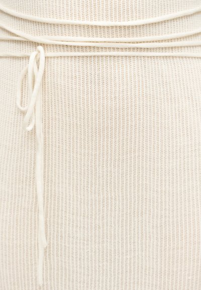 Gros plan sur un tissu tricoté côtelé beige avec un nœud coulissant noué sur le côté gauche, montrant la texture et le motif de couture.