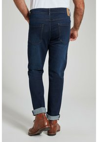 Jeans de mezclilla azul oscuro con corte ajustado, que cuentan con bolsillos traseros, puños enrollados y zapatos de cuero marrón. Tejido texturizado con ligero desgastado.