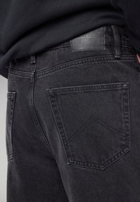 Musta farkut, joissa on suora lahkeensuukuvio, näkyvät ompeleet ja nahkapäällinen vyötäröllä, jossa lukee "AMERIKKA NYT."