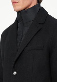 Homme portant un manteau en laine noir avec une doudoune noire à col montant zippée en dessous, se concentrant sur le haut du torse et la zone du cou.