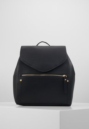 Schwarzer Leder-Rucksack mit vorderer Reißverschlusstasche, Klappenverschluss und kleinem Tragegriff oben, auf einem weißen Sockel vor grauem Hintergrund platziert.