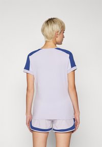 Camiseta deportiva de color púrpura claro con toques azules en los hombros y las mangas, combinada con pantalones cortos grises claros con ribetes azules. Textura suave.