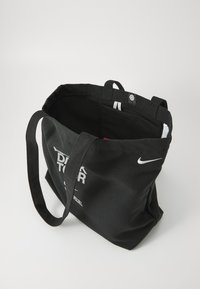 Black canvas tote bag with dual black handles, featuring white branding text and a small white logo on the inside. Simple, spacious design.