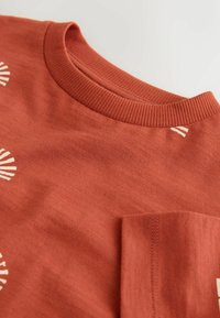 Rustfarvet t-shirt med en ribbet halsudskæring, der har en lomme og et skalsmotiv trykt i en lysere farve. Blødt, tekstureret stof.