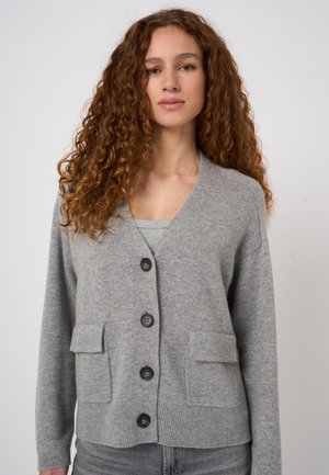 Woman with long curly hair wearing a light grey buttoned cardigan with two front pockets over a grey top.