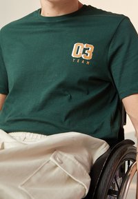 Zelené bavlněné tričko s kulatým výstřihem, s grafickým designem v oranžové a bílé barvě s nápisem "03 TEAM" na levé straně hrudi, hladká textura.