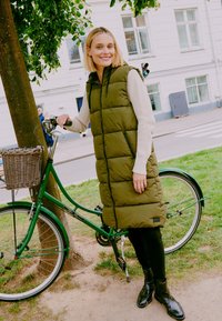 Olivgrüne Steppweste mit Kapuze, ausgestattet mit einem vertikalen Reißverschluss und gestepptem Design, getragen über einem cremefarbenen Pullover, kombiniert mit schwarzen Leggings und Stiefeln.