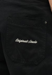 Τσέπη μαύρου τζιν παντελονιού με λευκή κεντημένη επιγραφή «Originals Studio» και μεταλλικό τρουκ στην επάνω γωνία.