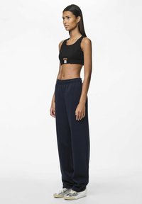 Zwarte cropped top met uitsnede aan de voorkant, gecombineerd met loszittende marineblauwe sweatpants en zilveren sneakers, staand op een effen achtergrond.