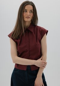 Koszulka z krótkim rękawem w kolorze burgundowym, zapinana na guziki, z wyostrzonym kołnierzykiem, wykonana z gładkiego materiału, zestawiona z ciemnymi dżinsami. Prosty design, brak wzorów.