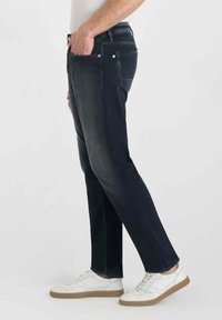 Jeans en denim foncé à coupe droite, avec un design classique à cinq poches et une légère décoloration. Portés avec des baskets blanches.