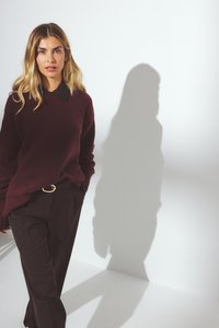 Bordo adīta džemperis ar melnu apkakli; apvienots ar melnām biksēm. Audums izskatās mīksts, ar vaļīgām piedurknēm un brīvu siluetu.