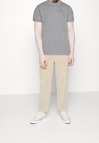 Muž v šedém polokošili s krátkým rukávem, béžových kordových kalhotách a bílých vysokých teniskách stojí před jednobarevným bílým pozadím.