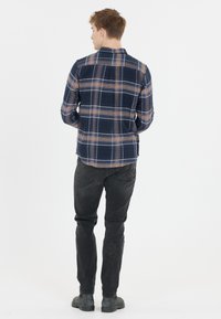 Miesten tummansininen ruudullinen flanellipaita, jossa on ruskean ja beigen sävyjä. Pehmeä rakenne, napitettava muotoilu ja kaareva helma. Yhdistetty mustiin housuihin.