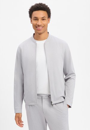 Junger Mann lächelt, trägt eine hellgraue Jacke mit Reißverschluss über einem weißen Hemd und passende hellgraue Hosen, steht vor weißem Hintergrund.