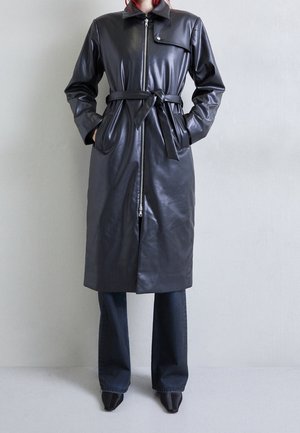 Persona con un abrigo de cuero negro hasta la rodilla con cinturón, cierre frontal con cremallera, manos en los bolsillos, combinado con pantalones oscuros y zapatos negros puntiagudos.