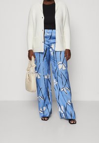 Persona con pantalones anchos de flores azules, camiseta negra, cárdigan blanco, sandalias negras y sosteniendo un bolso beige frente a un fondo liso.