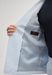 Main tenant un blazer bleu clair ouvert montrant une doublure intérieure subtilement imprimée et une poche intérieure avec une étiquette de marque.