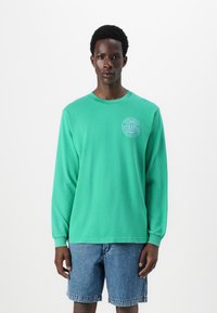 Long-sleeve green cotton t-shirt with a round neckline, featuring a white circular graphic on the chest; paired with denim shorts.