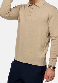 Camisa polo de manga larga beige con cuello, tapeta de tres botones, puños acanalados y dobladillo. Textura suave y diseño ajustado, combinada con pantalones azul marino.