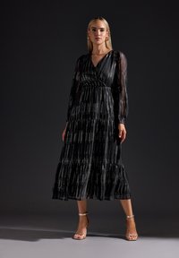 Svart klänning med långa genomskinliga ärmar, flerlagersdesign och metalliska vertikala ränder. Har en V-ringning och en svängig silhuett. Bärs med snabblänkade klackar.