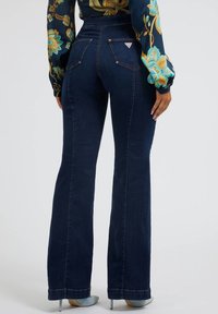 Kvinde iført mørkeblå højtaljede flared jeans, en langærmet mønstret bluse og lyseblå hæle, stående foran en ensfarvet baggrund.