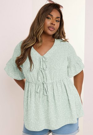 Kvinde iført lysegrøn blomsterprintet kortærmet bluse med knuder foran og blå cowboyshorts, poserer mod en lys baggrund.