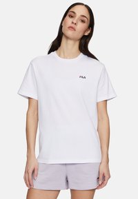 La maglietta bianca in cotone presenta maniche corte e un collo a giro, con un piccolo logo scuro sul petto sinistro. Indossata con pantaloni corti lilla.