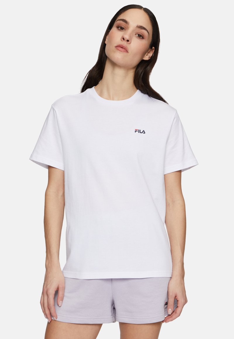 La maglietta bianca in cotone presenta maniche corte e un collo a giro, con un piccolo logo scuro sul petto sinistro. Indossata con pantaloni corti lilla.
