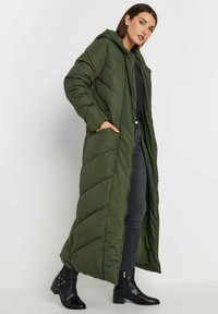 Długa oliwkowozielona kurtka puchowa z kapturem, z fasonem w romby, bocznymi kieszeniami i pełnej długości. Nosi pod nią czarną bluzkę i dżinsy.