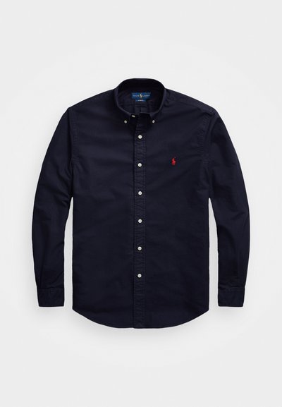 Temno modra srajca z gumbi in s koničastim ovratnikom, dolgimi rokavi in majhnim rdečim logotipom na prsih. Bombažni material z gladko teksturo.