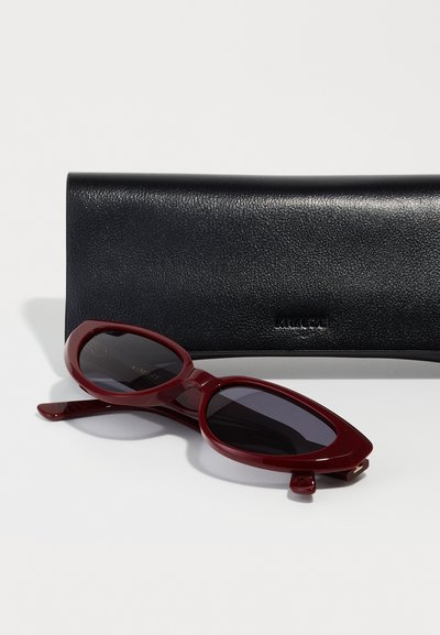 Red cat-eye sunglasses with dark lenses, resting beside a textured black leather case, featuring a subtle embossed logo.