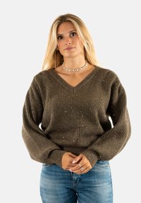 Maglione a costine verde oliva con scollo a V, maniche lunghe e dettagli in filo metallico. Abbinato a jeans blu chiaro e una collana a conchiglia.