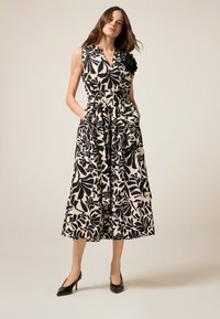Ärmelloses Kleid in einem schwarzen und cremefarbenen Blumenmuster, mit V-Ausschnitt, Taschen und einem Stoffblumenakzent an der Schulter.