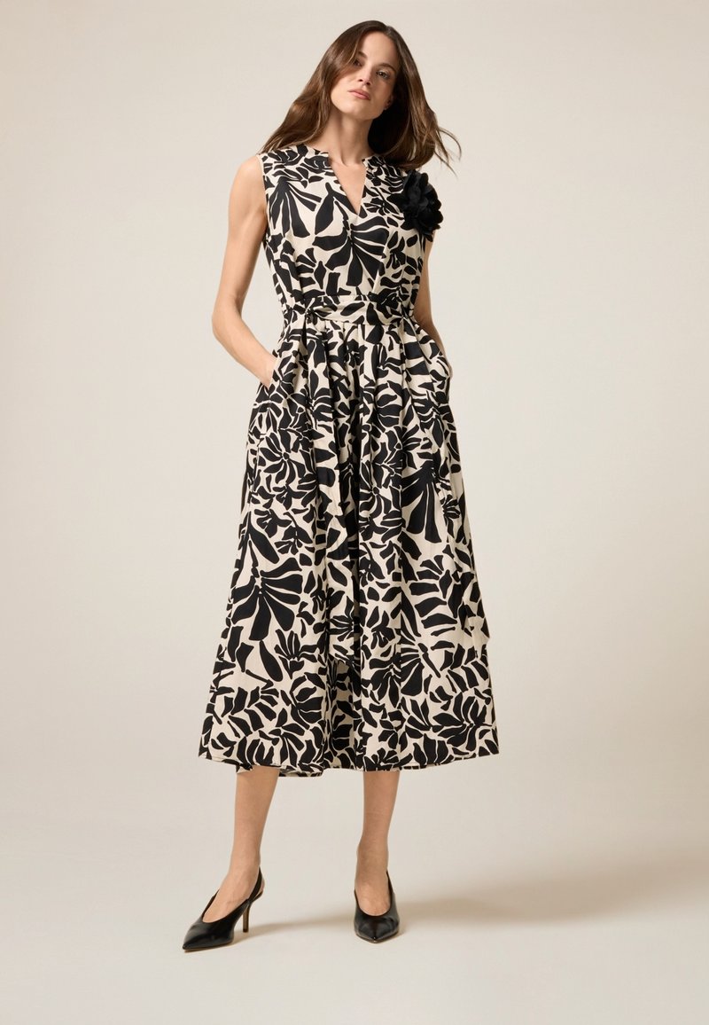 Ärmelloses Kleid in einem schwarzen und cremefarbenen Blumenmuster, mit V-Ausschnitt, Taschen und einem Stoffblumenakzent an der Schulter.