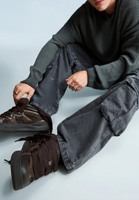 Sötétzöld bordázott pulóvert, szürke cargo nadrágot és fekete, túlságosan nagy fűzőjű tornacipőt viselő személy ül a padlón, és egy tornacipőt tart a kezében.