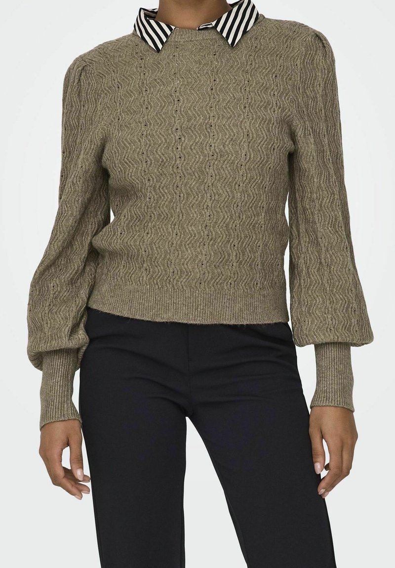 Femme portant un pull en maille texturée taupe avec des manches ballon et une chemise à col rayée noir et blanc, associée à un pantalon noir.