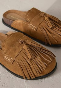 Zapatos de ante marrón tipo slip-on con un diseño redondeado, que presentan flecos decorativos, acentos de costura y una suela texturizada para mayor agarre.