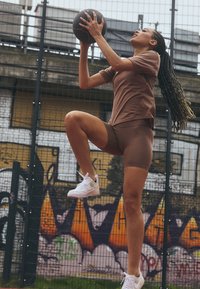 T-shirt de sport marron et shorts, baskets blanches, en train de sauter avec un ballon de basket devant un fond en graffiti texturé et coloré.