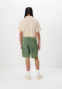 Beżowa koszula z krótkim rękawem, zielone szorty dżinsowe o wyblakłej fakturze i off-white sneakersy. Wyeksponowane z tyłu na białym tle.