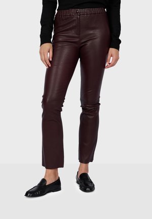 Pantalon en cuir marron foncé coupe slim porté avec des mocassins noirs et un top noir à manches longues, montré de la taille aux pieds sur un fond clair.