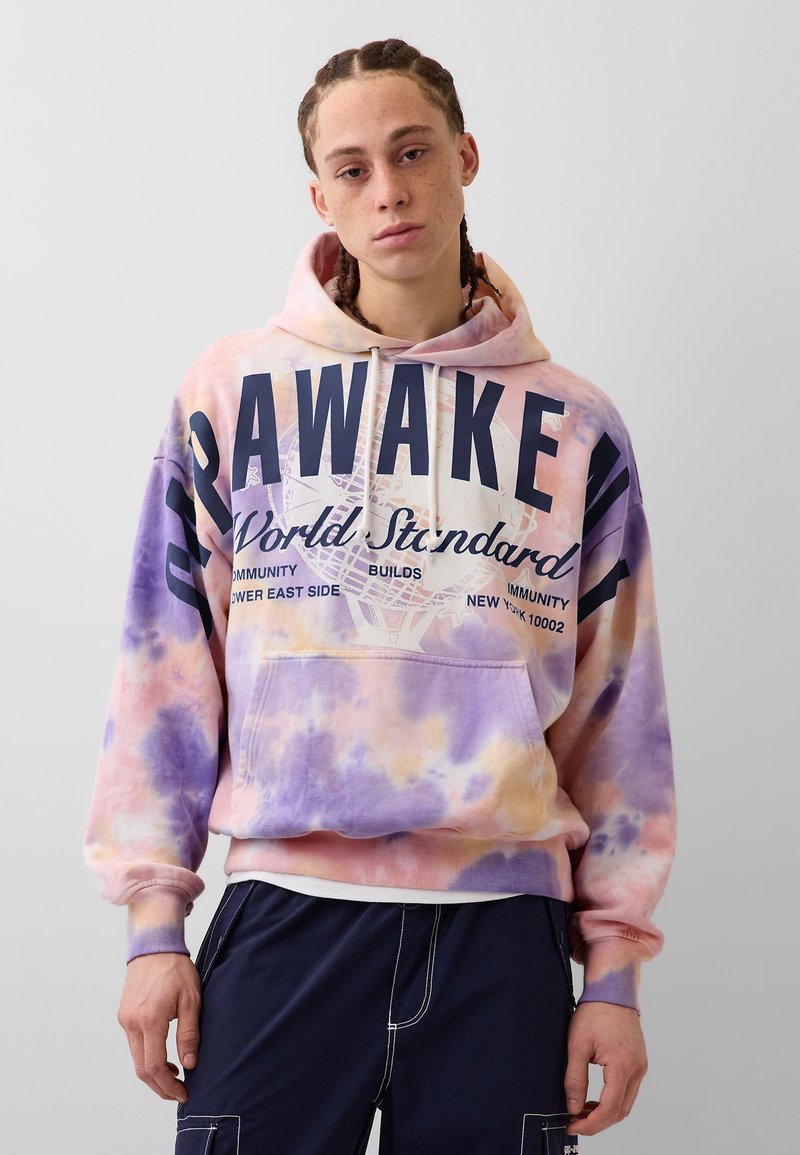 Άτομο με πλεξούδες, που φοράει παστέλ tie-dye φούτερ με μεγάλα μπλε γράμματα και μπλε παντελόνι, με απλό ανοιχτό φόντο.