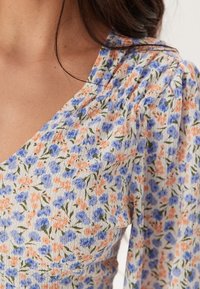 Tissu à motifs floraux avec de petites fleurs bleues et orange sur fond blanc, doté de détails texturés et plissés le long de l'encolure.