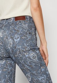 Gros plan sur une personne portant un jean bleu à motif cachemire et un haut beige, montrant la poche arrière et une partie du bras.