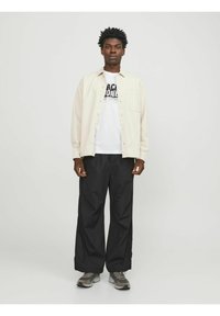 Camisa de botón beige clara sobre una camiseta blanca con texto gráfico negro, combinada con pantalones negros holgados y zapatillas grises.
