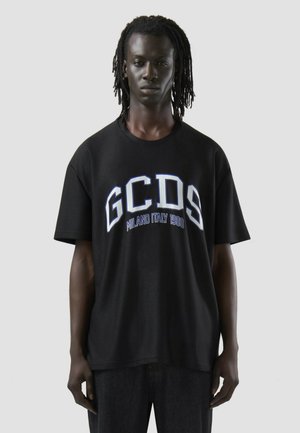 Mand med dreadlocks iført en sort oversized T-shirt med teksten "GCDS Milano Italy 1988" mod en ensfarvet lys grå baggrund.