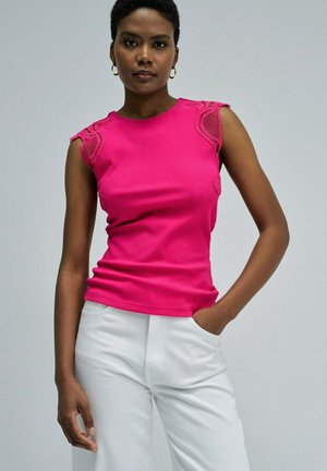 Mujer de pie con una mano en el bolsillo, vistiendo un top sin mangas de color rosa brillante con detalles de encaje y pantalones blancos, fondo neutro.