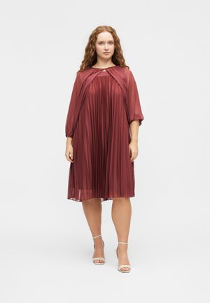 Žena s dlhými kučeravými vlasmi, oblečená v plisovaných bordových šatách po kolená s priehľadnými rukávmi a béžovými členkovými topánkami na opätku, stojaca na bielom pozadí.